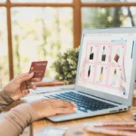 Une personne devant un ordinateur connecté sur un site de vente en ligne tient une carte bancaire à la main.
