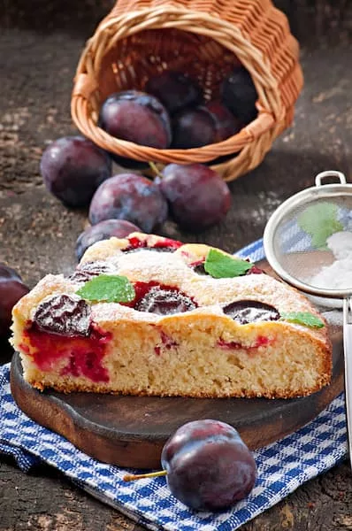 Gâteau aux prunes fraîches saupoudré de sucre glace avec fruits frais et passoire décorative sur torchon à carreaux bleus